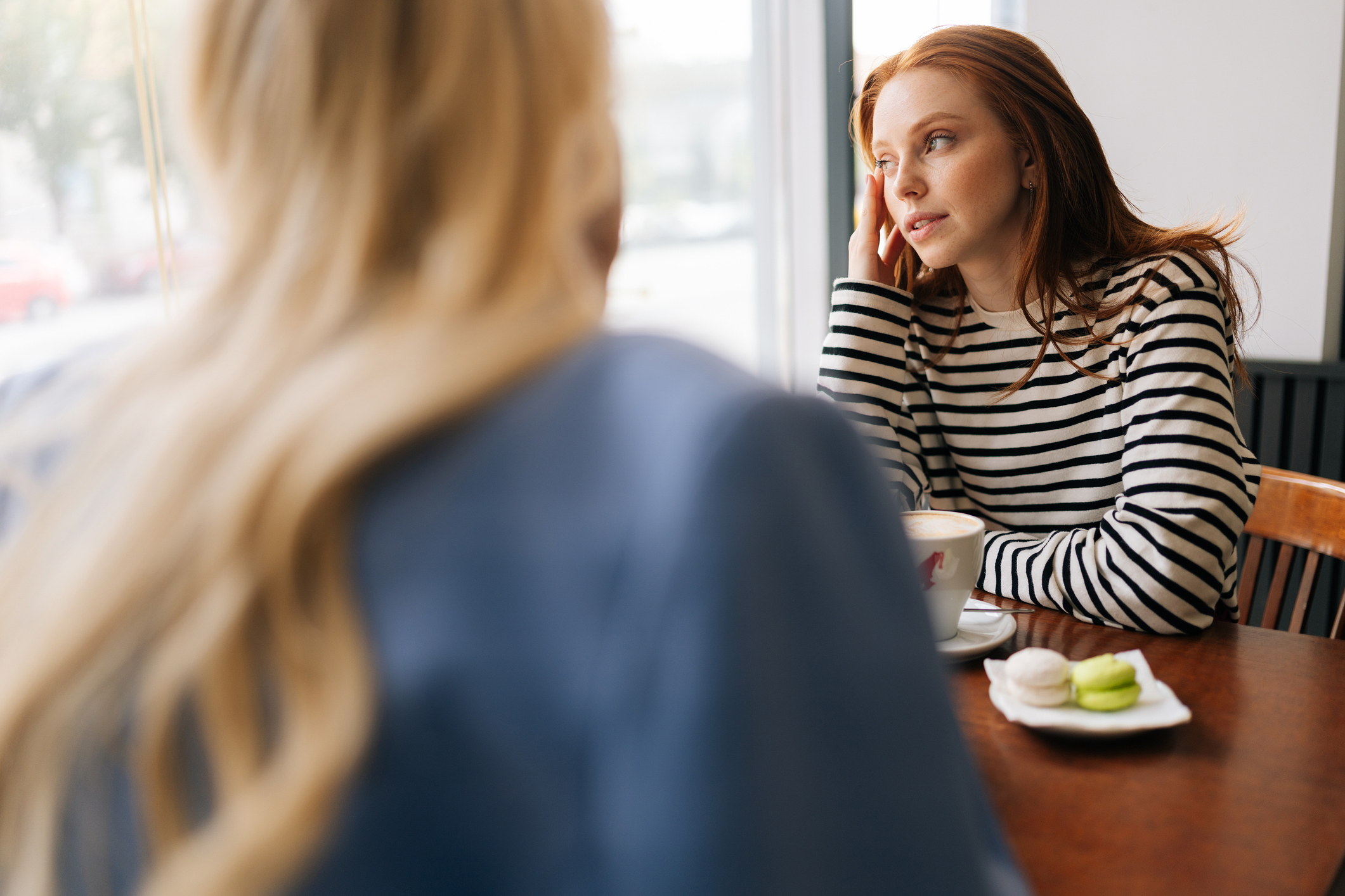 Rear view of unrecognizable blonde female talking to pensive woman friend, spending time together during drinking coffee at cozy cafe sitting at table by window